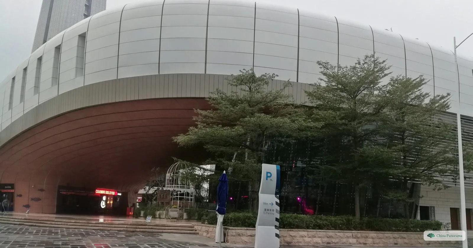 The white, curved front of the Sea World Culture and Arts Center in Shekou on a grey morning, with a dark red underside, young trees and a wet forecourt