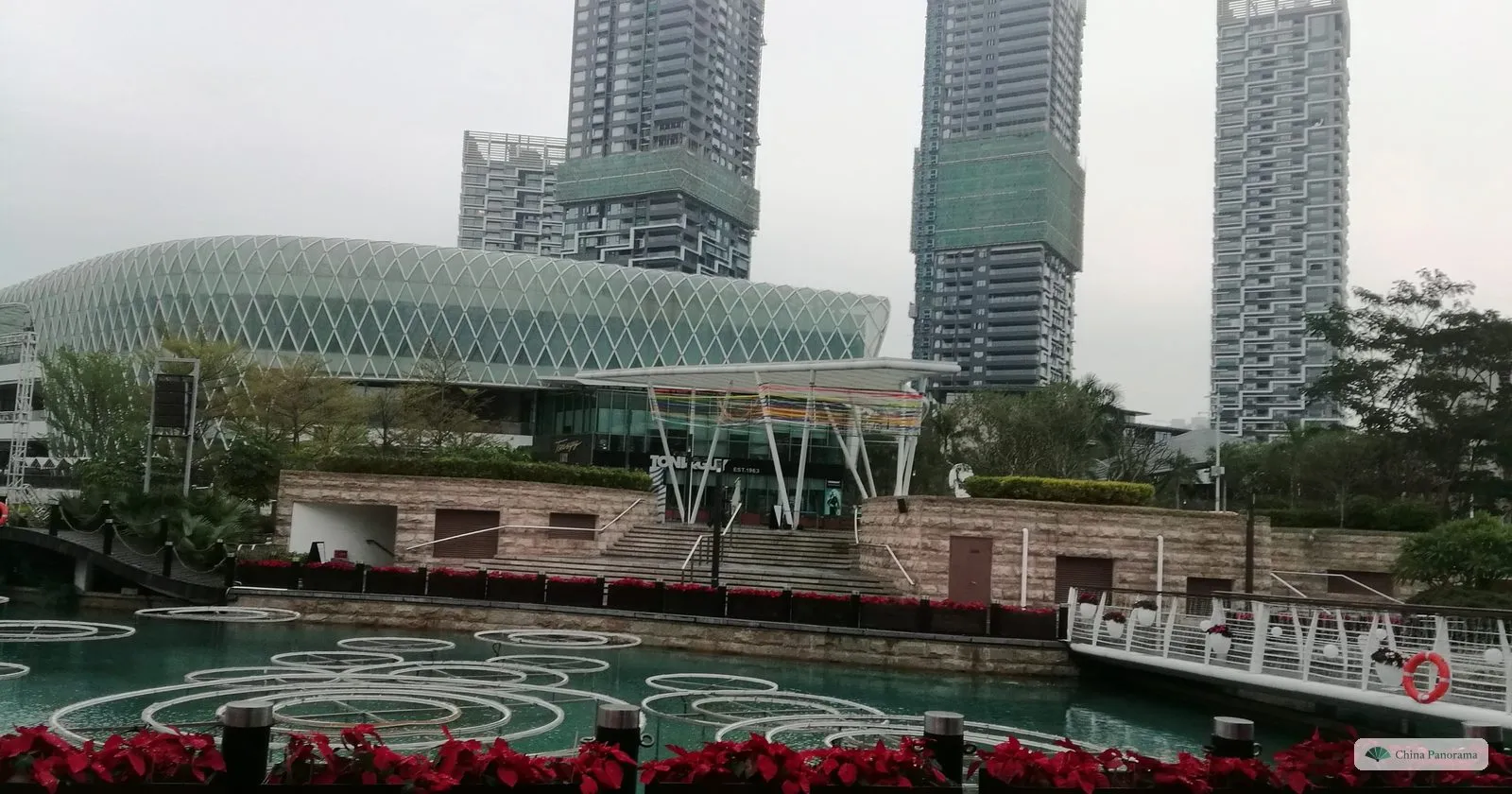 The lagoon at Sea World in Shekou with a swirling pattern on the water, red poinsettias along the edge, a glass-domed building and three tall residential towers behind