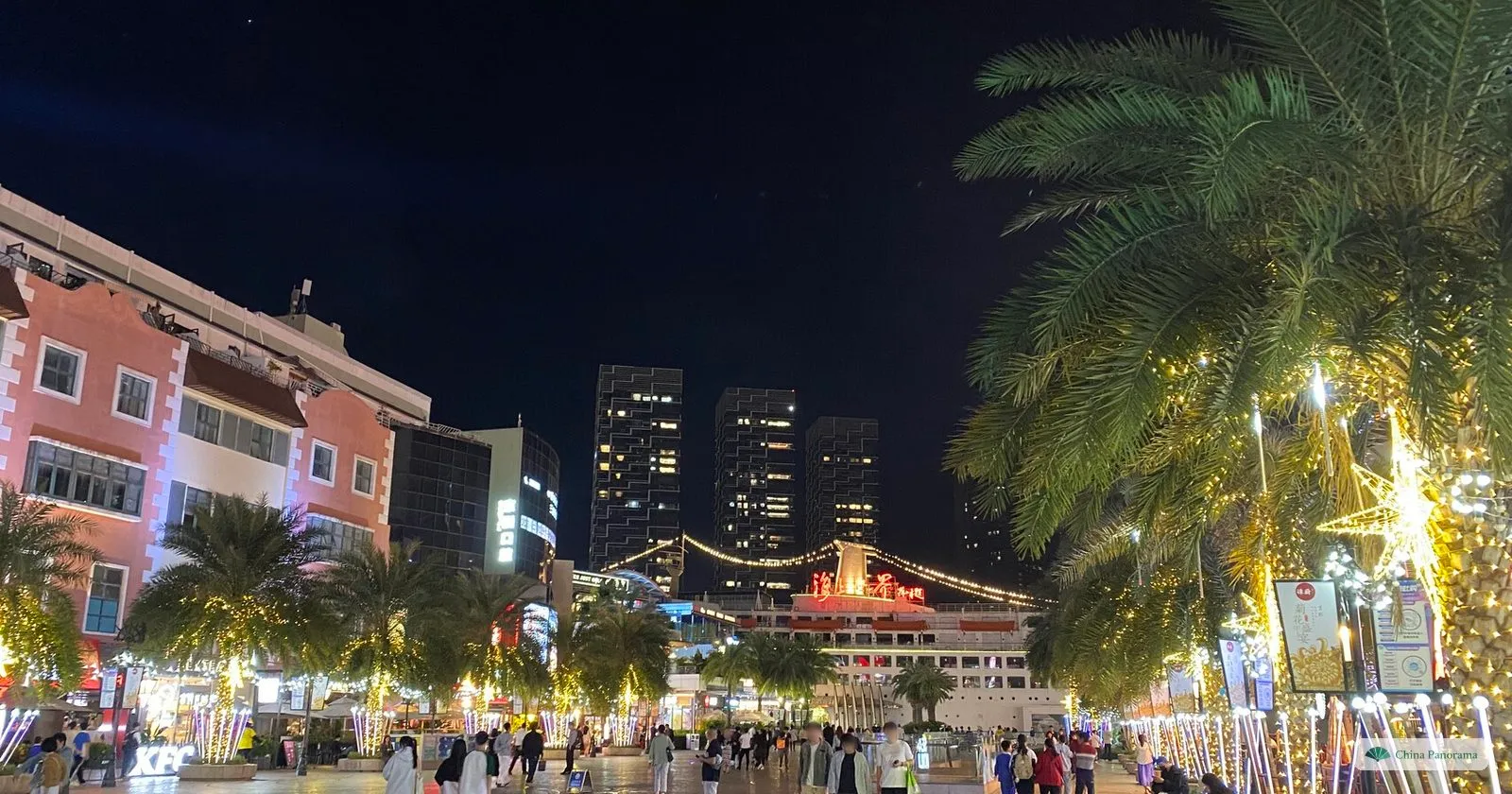 The Sea World plaza in Shekou at night, crowded with people, lit palm trees on the right and the illuminated Minghua at the far end of the square