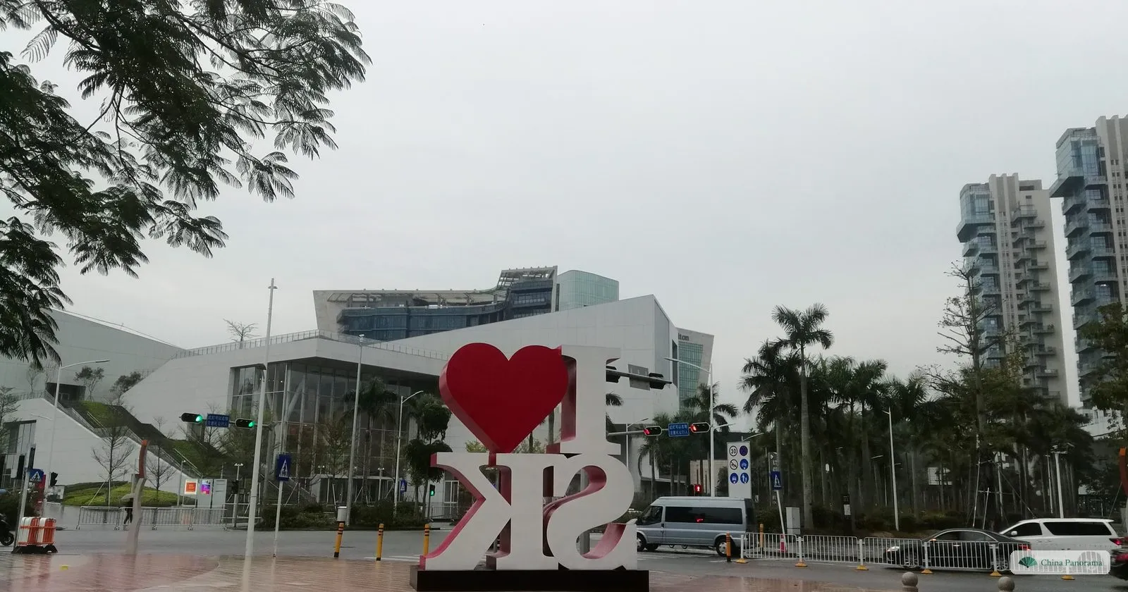 A large red-and-white sign reading I ♥ SK on a wet plaza in Shekou, with modern white buildings, palm trees and residential towers behind