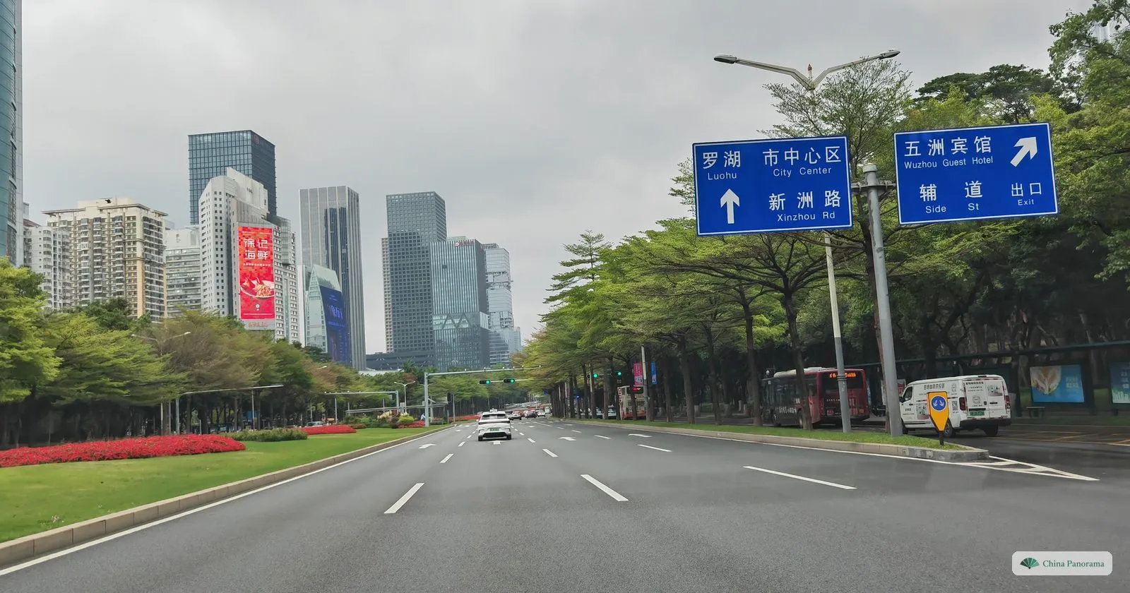 Blue road signs over Shennan Boulevard in Chinese and English: Luohu and City Center straight on via Xinzhou Rd, Wuzhou Guest Hotel to the right by the side street exit