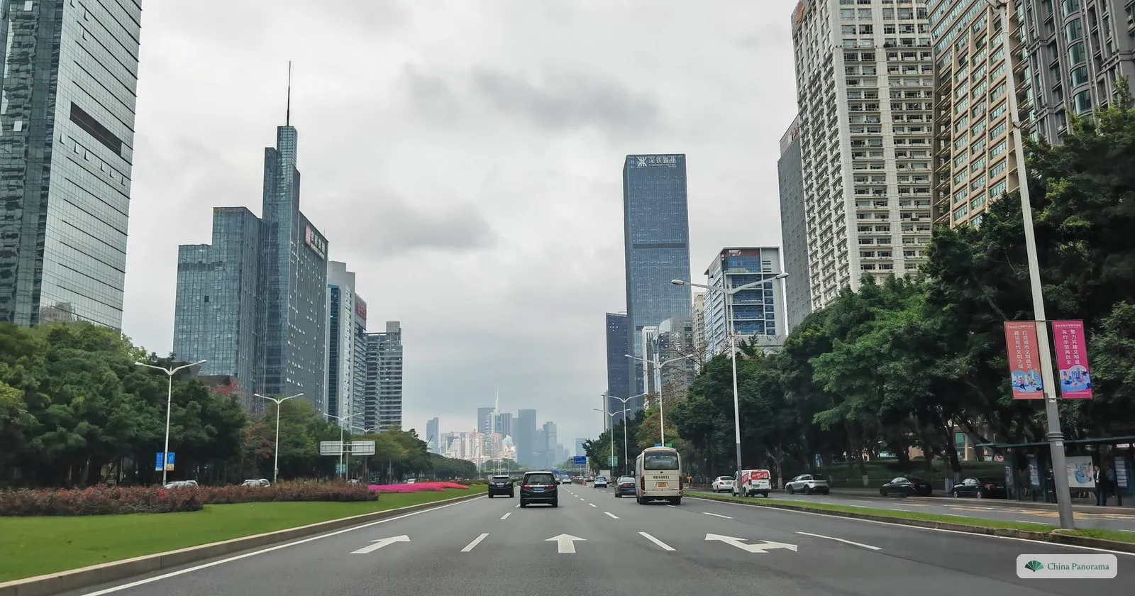 A row of glass office towers along Shennan Boulevard, one with a tall spire, at the end of a wide road with lane arrows and a green verge