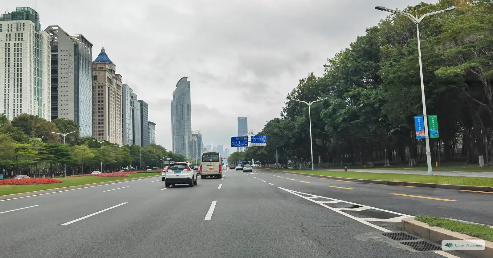Shennan Boulevard under grey cloud: a wide, almost empty road between planted strips with red flowers, office buildings on the left and a line of trees on the right
