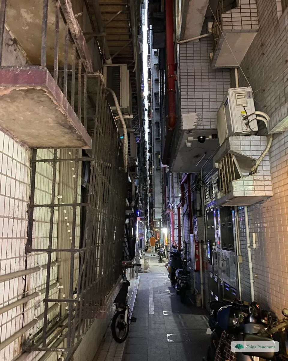 A very narrow lane between apartment blocks at dusk, barred windows and air-conditioning units on both sides, scooters parked along one wall