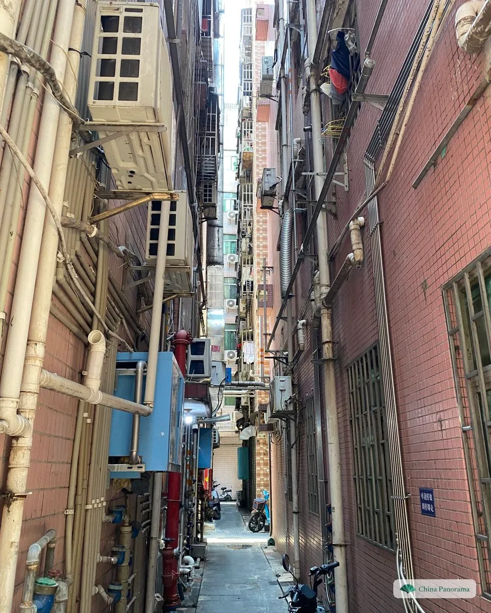 A narrow alley between tall tiled apartment blocks in an urban village, air-conditioners and pipes on the walls and a strip of sky far above