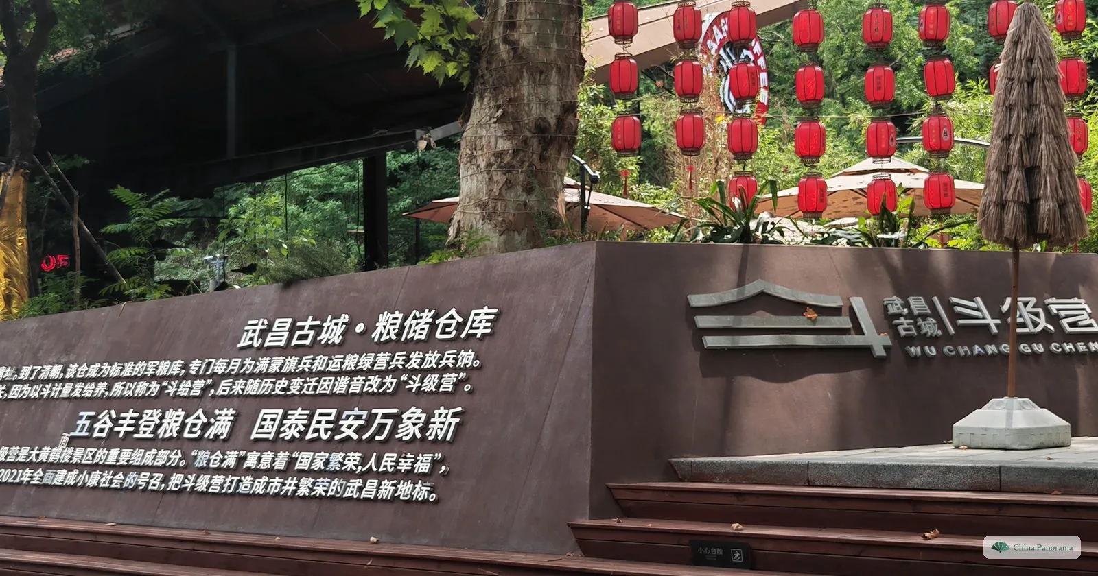 A dark brown wall with white characters and a logo, beneath trees hung with rows of red lanterns