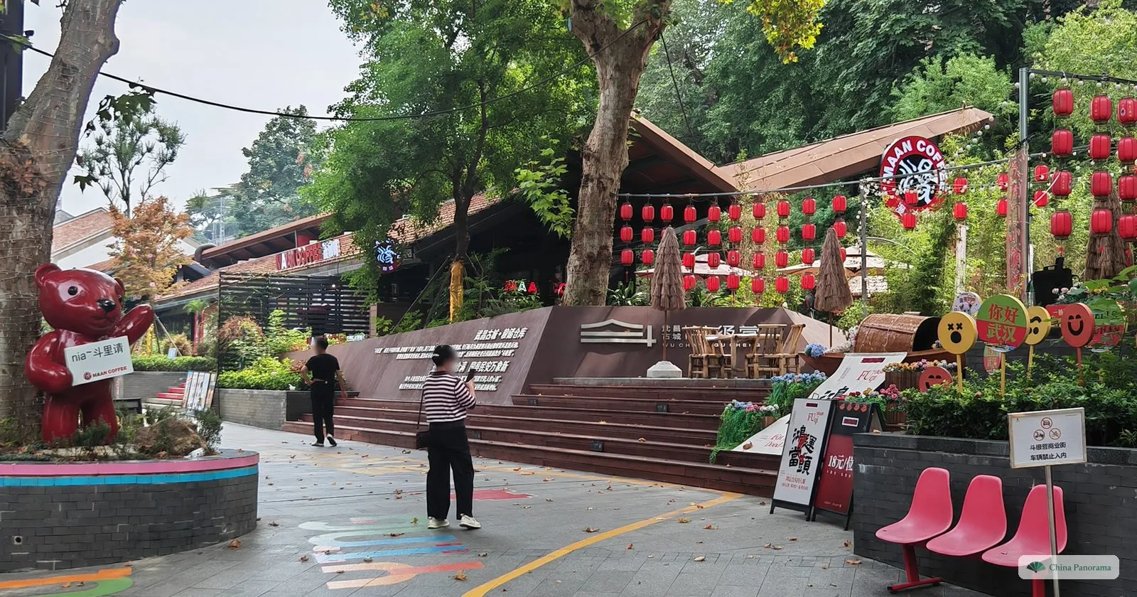 A small square with a red teddy-bear figure, steps up to a café terrace hung with red lanterns, two people walking