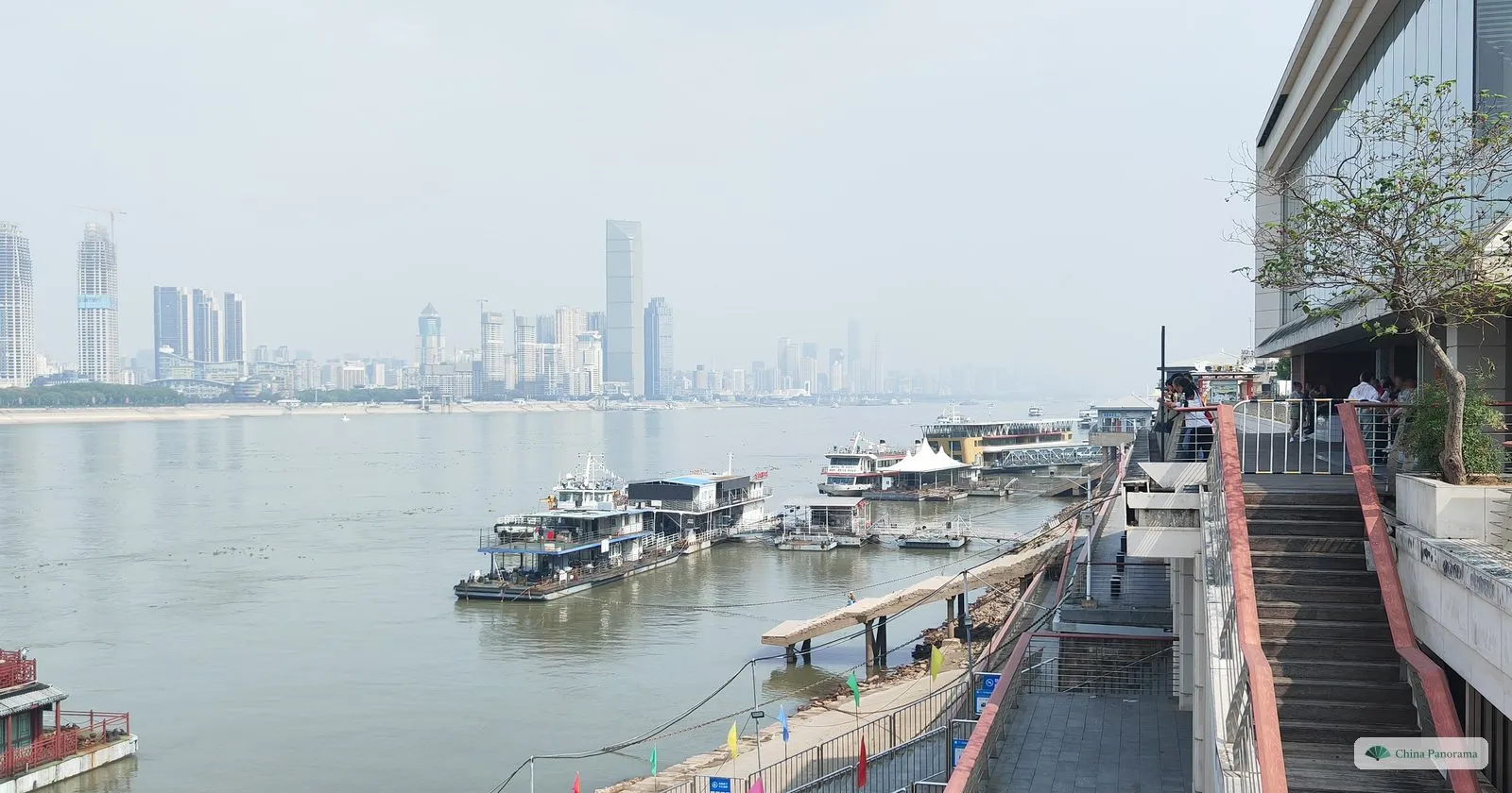Looking down from a riverside walkway at floating piers and moored boats on a wide river, a skyline of towers on the far bank