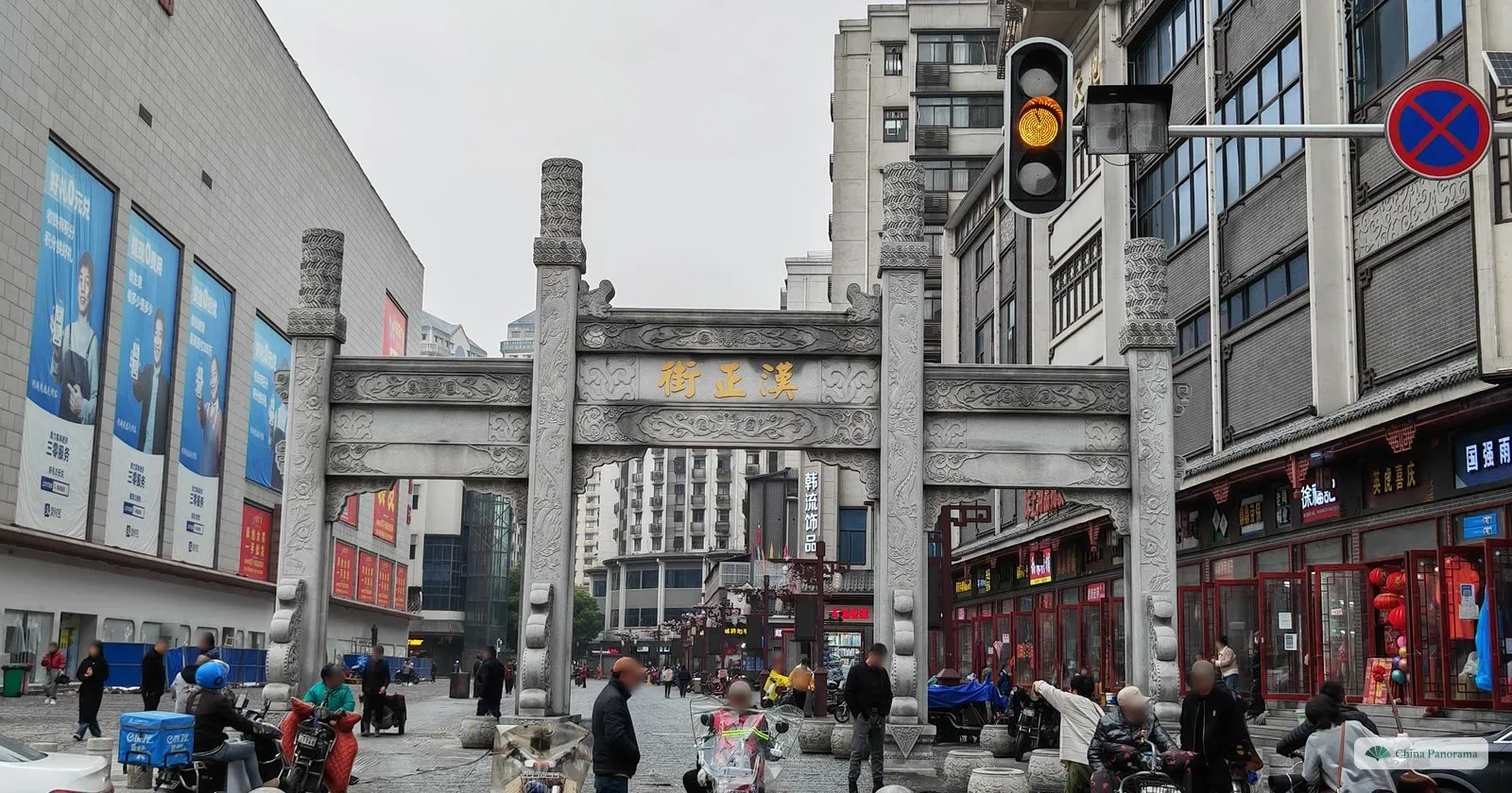 A grey stone archway with carved pillars and three gold characters on its crossbeam, on a street with modern shops, a traffic light and pedestrians