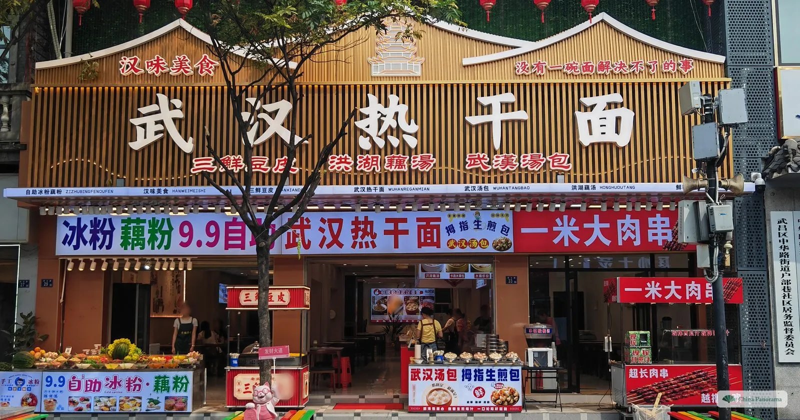 A restaurant front with large white characters 武汉热干面 on a slatted wooden facade, smaller signs for doupi, lotus-root soup and soup buns, and food counters below