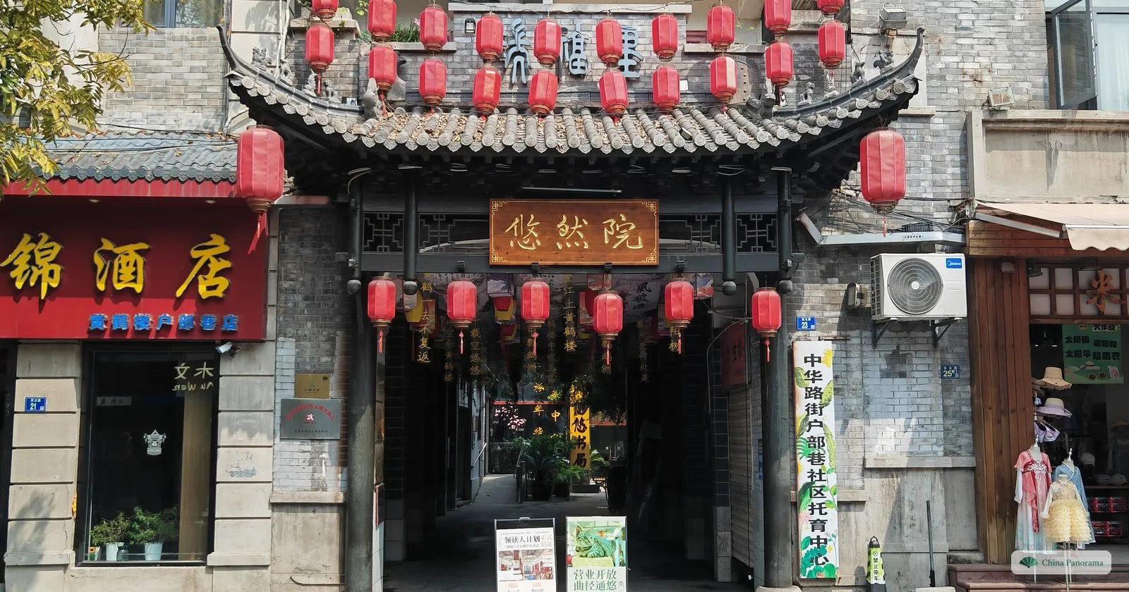 A grey-brick gate with a carved wooden canopy hung with red lanterns and a plaque 悠然院, a shop sign beside it