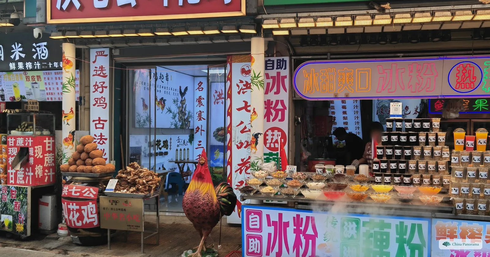 A snack stall with tubs of toppings for ice jelly, a model rooster and handwritten signs for sugar-cane juice and beggar’s chicken