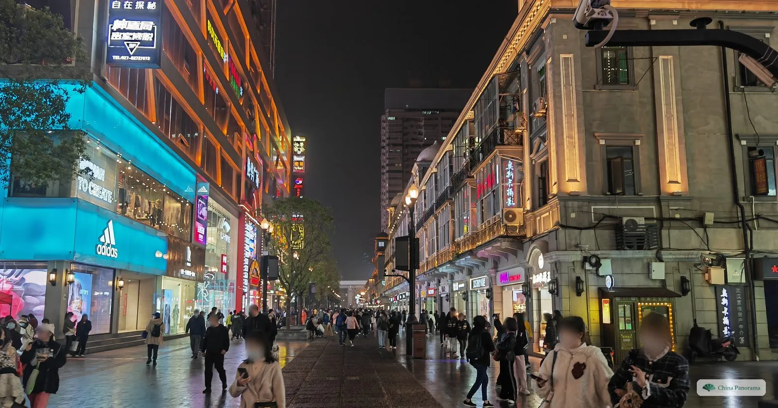 A wide pedestrian street at night between brightly lit shopfronts and a row of older buildings with balconies, people walking on wet paving
