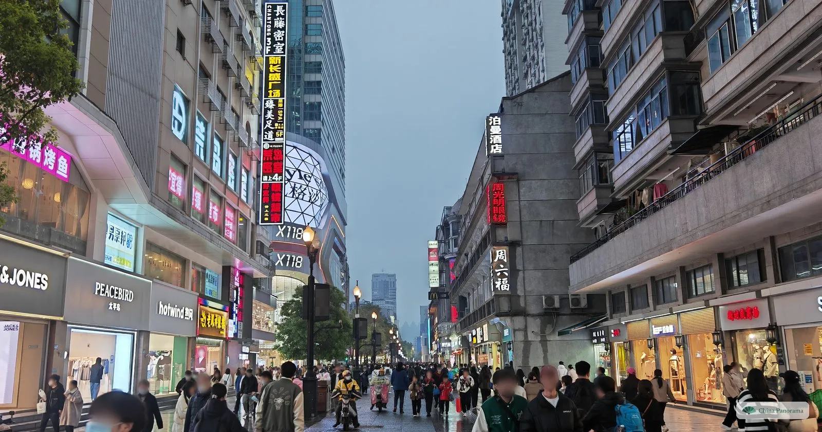 A pedestrian shopping street at dusk lined with shops and old buildings with balconies, crowds walking under the street lamps