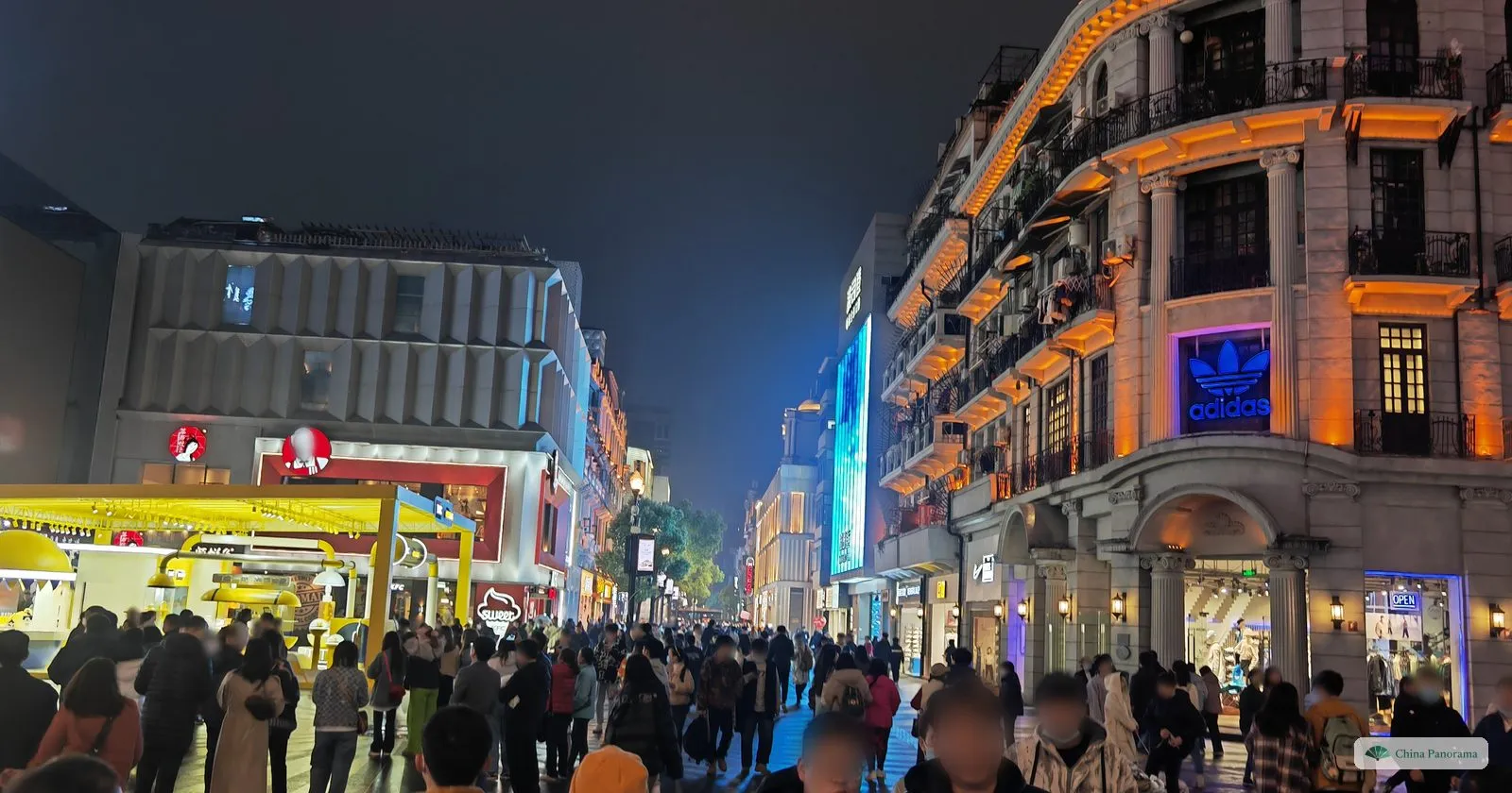 A crowded pedestrian street at night beside a cream-coloured building with columns and an illuminated sign