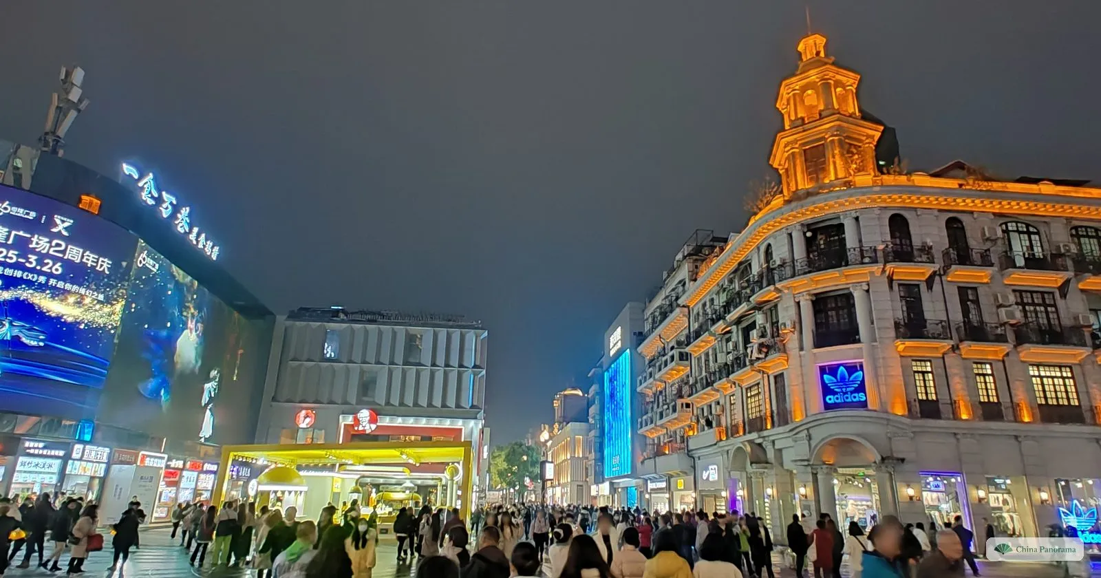 An ornate corner building with a gold-lit tower and balconies at night, a giant video screen on the left and a crowd in the square