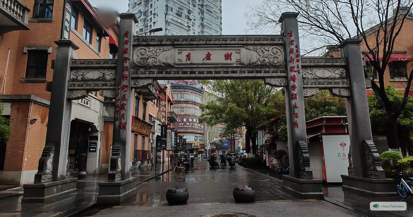 A stone archway with red calligraphy on its pillars and a carved crossbeam, over a wet street lined with red-brick buildings
