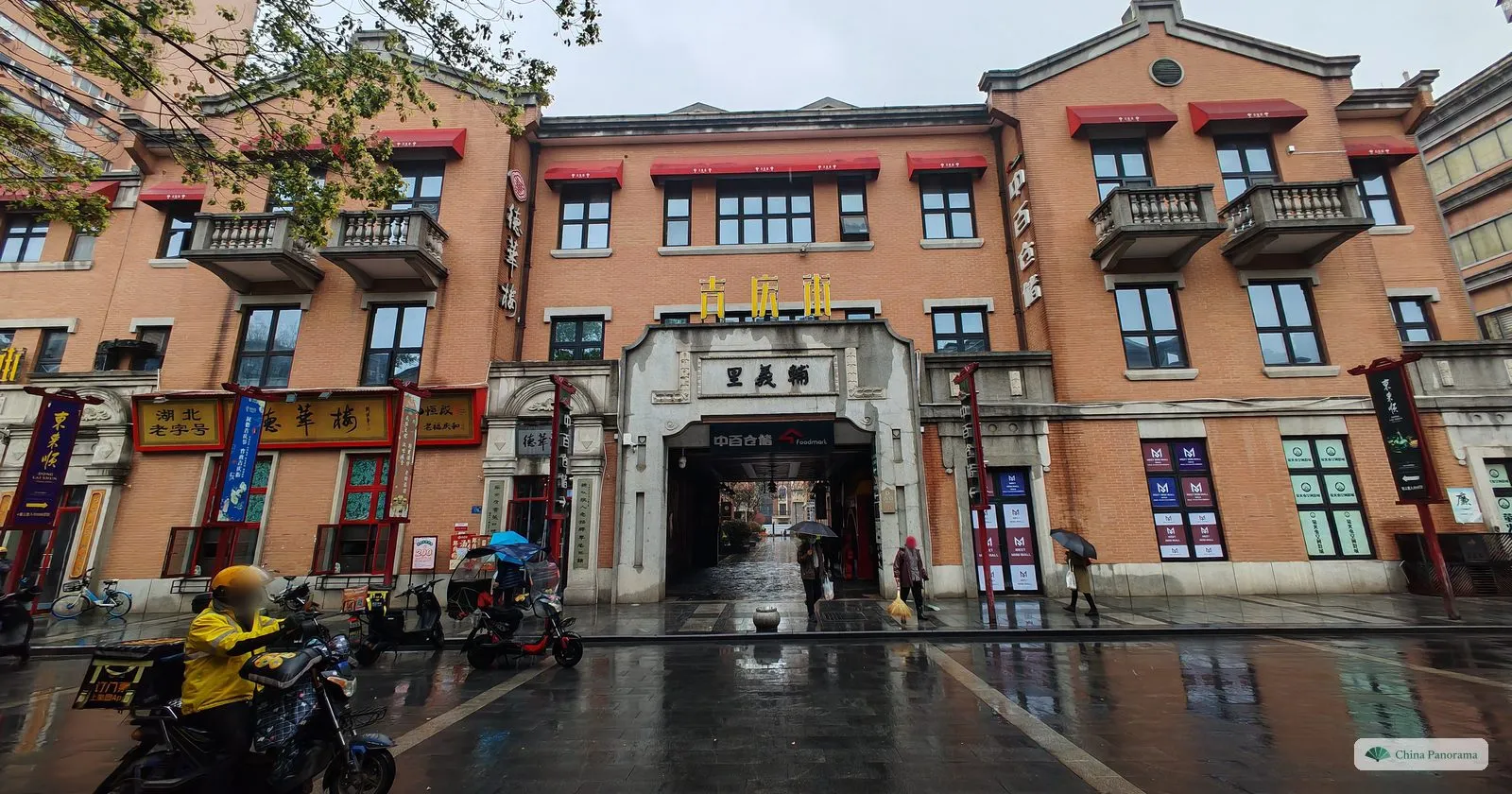 A red-brick building with white balconies and a stone gateway in the middle, a delivery rider passing on a wet street