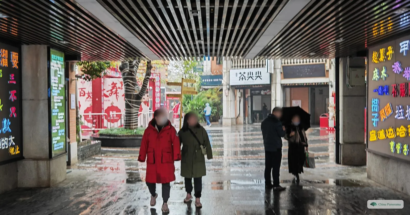 A covered passage with walls of neon characters, two people in red and green coats walking in the rain