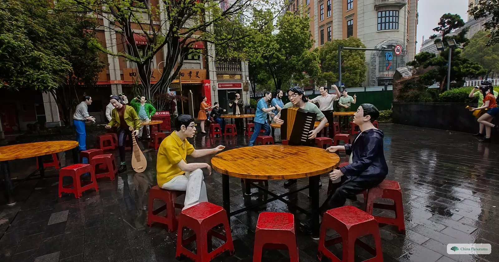 Life-size coloured figures of men playing an accordion, a pipa and cards round a table with red plastic stools, on a wet square