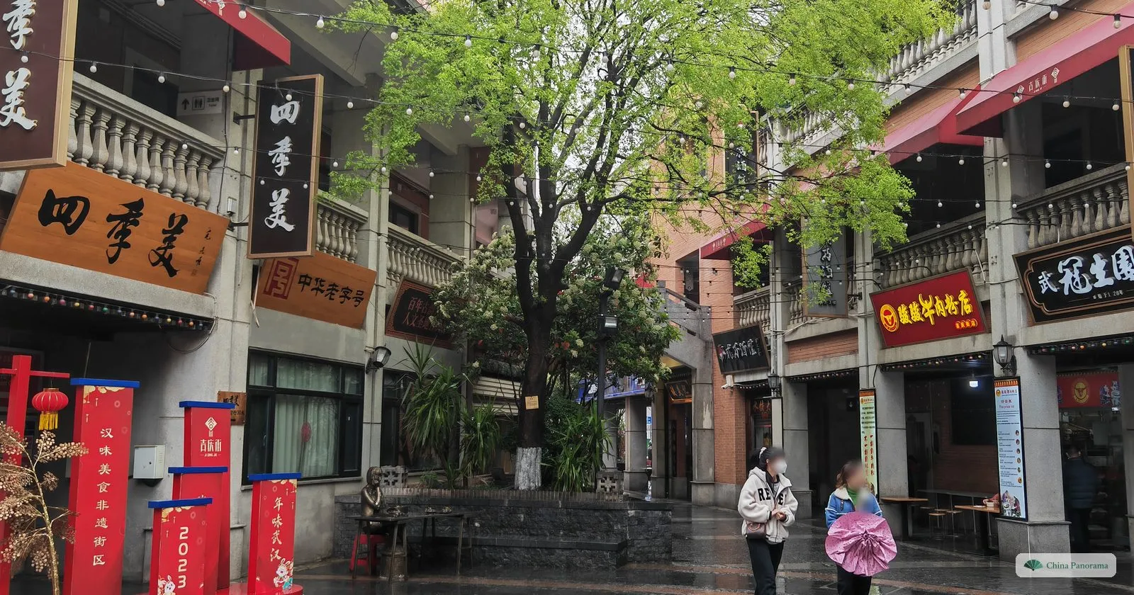 A lane of red-brick shops with wooden signboards 四季美, a tree in fresh leaf and two people with umbrellas