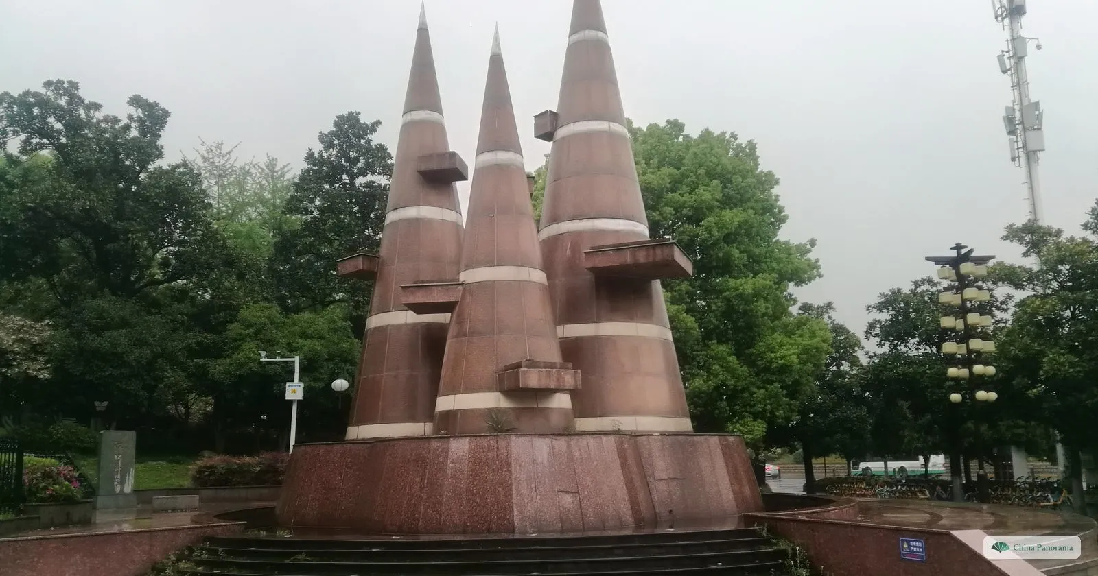 A monument of three tall granite cones with white bands on a round red stone base, trees around it on a rainy day