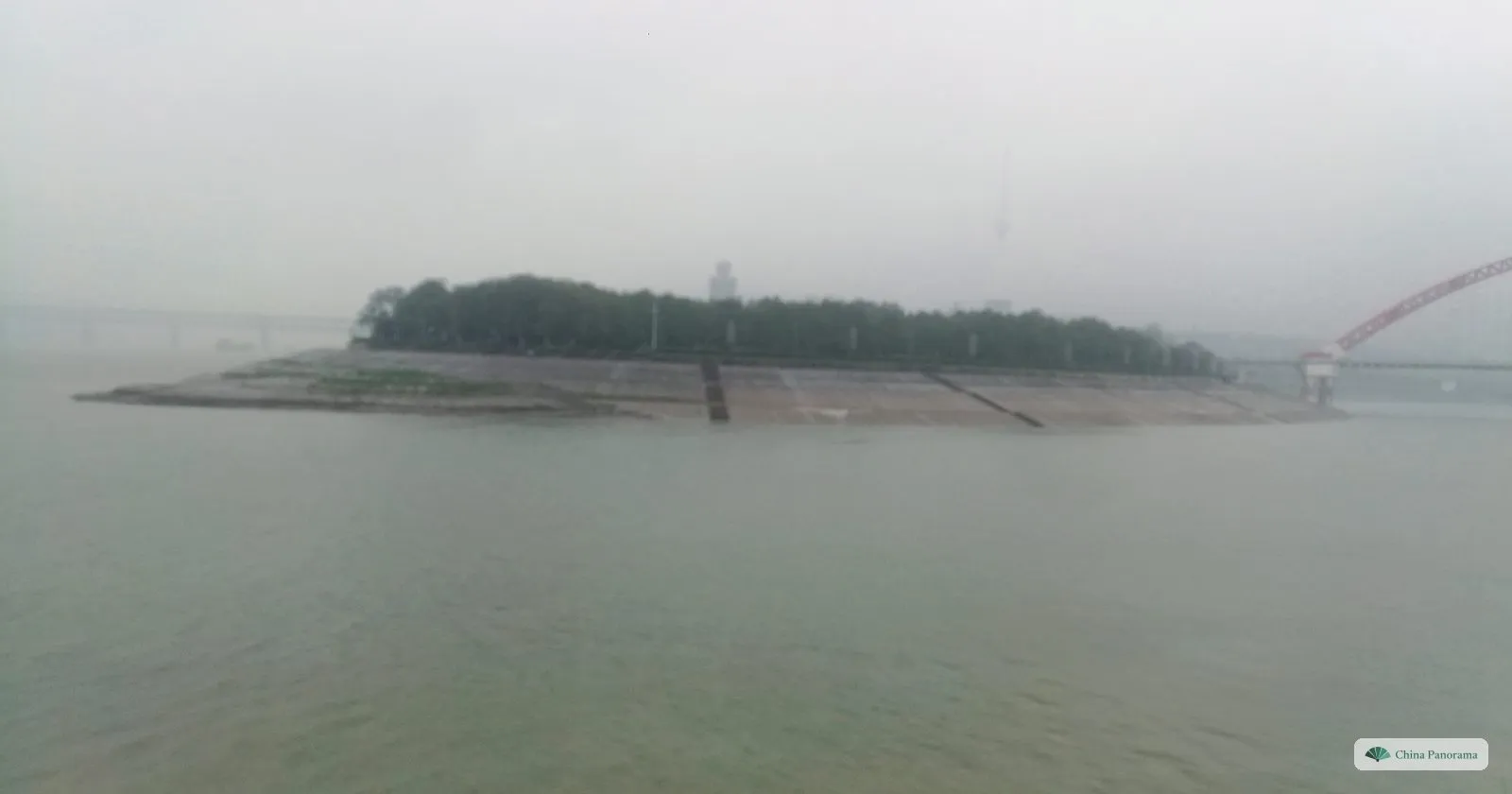 A low wooded spit of land with a sloping stone embankment in a misty river, the red arch of a bridge at the right edge