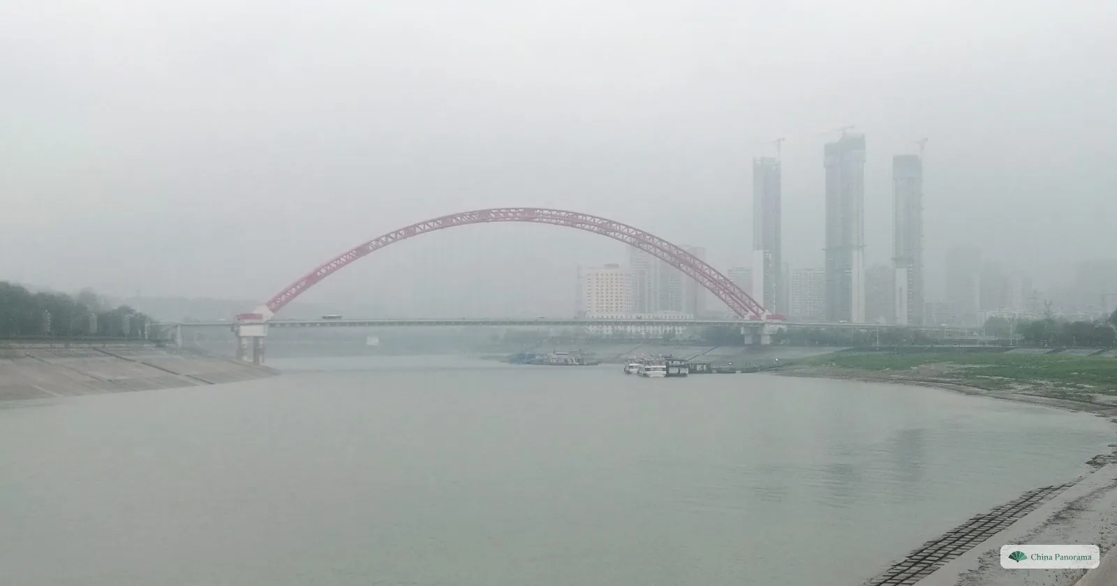 A red steel arch bridge over a grey river in rain and mist, tall towers half hidden behind it
