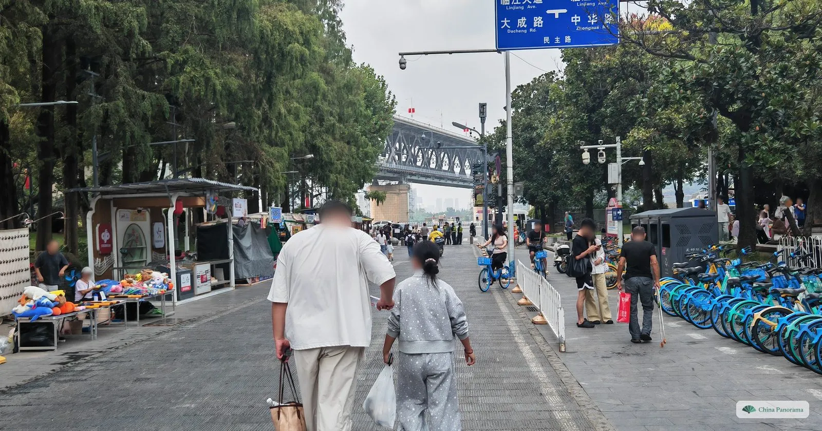 A street with souvenir stalls and rows of blue share bikes leading towards a steel bridge, a blue road sign overhead