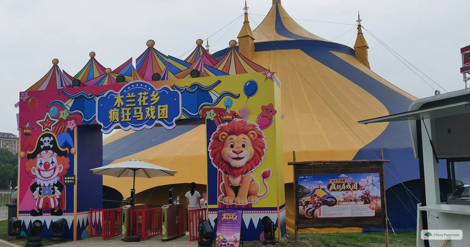 A circus entrance with a cartoon lion and clown and the words 木兰花乡 疯狂马戏团, a yellow and blue big top behind