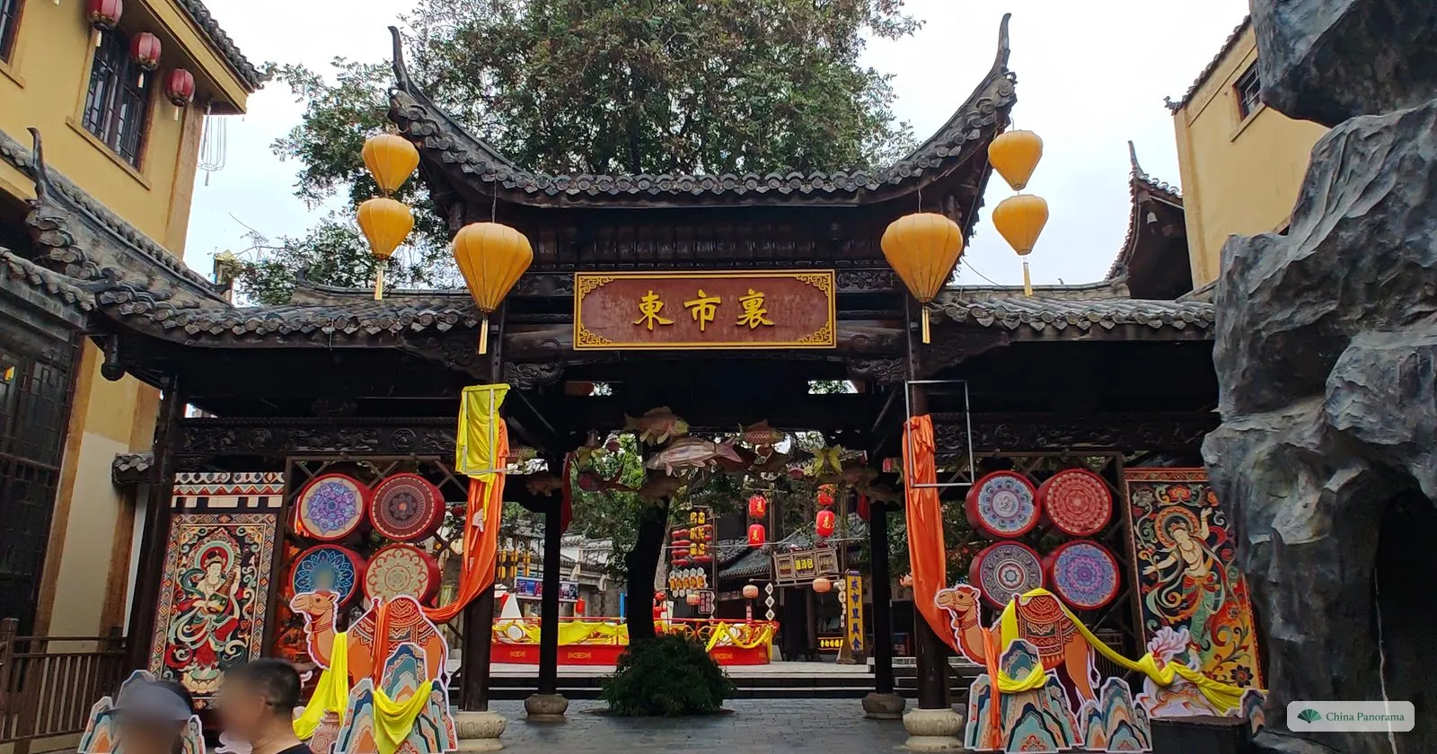 A dark wooden gateway with a plaque 東市裏, painted panels, camel cut-outs and orange lanterns