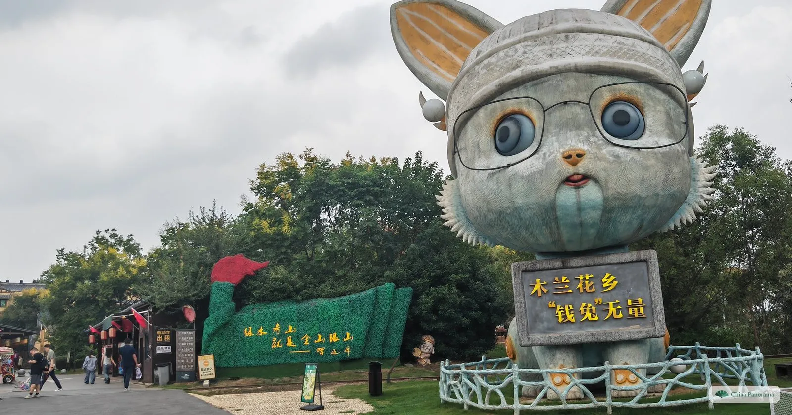A giant statue of a rabbit in glasses and a cap holding a sign, a hedge sculpture of a bird behind
