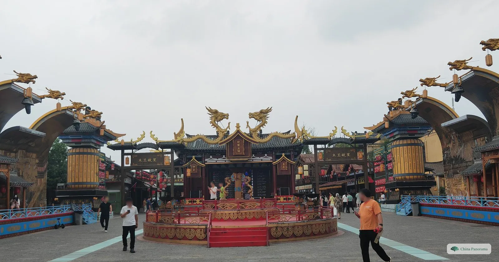 A wide square ringed by fantasy buildings with golden dragons on their roofs and drum-shaped towers, a round red stage in the middle