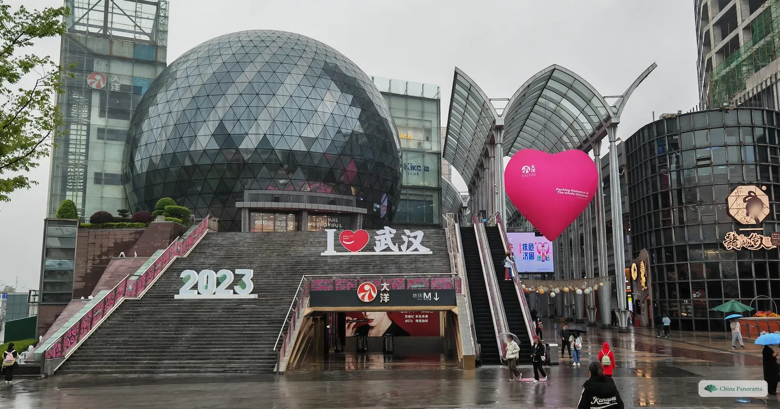 A shopping street with a glass dome, a wide stairway with the letters I ♥ and 2023, a pink heart balloon and people with umbrellas