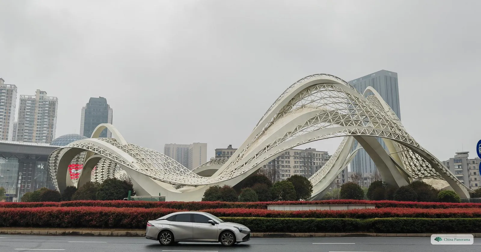 A white steel lattice sculpture of sweeping arches over a roundabout planted with red shrubs, a car passing in the rain