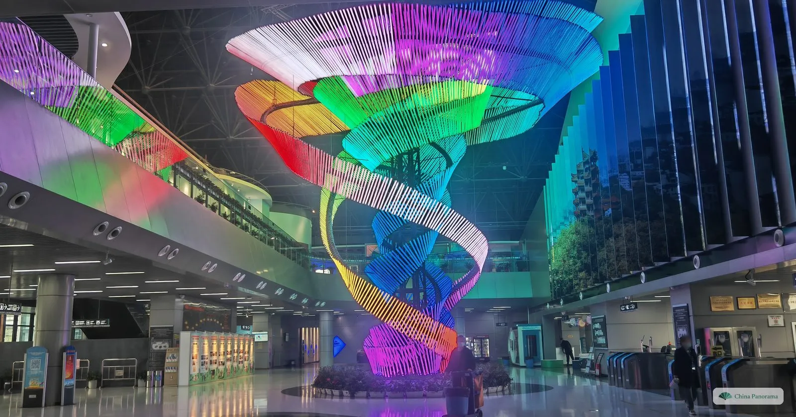 An entrance hall with a tall double helix of rainbow-coloured light tubes rising through several floors, ticket gates on the right