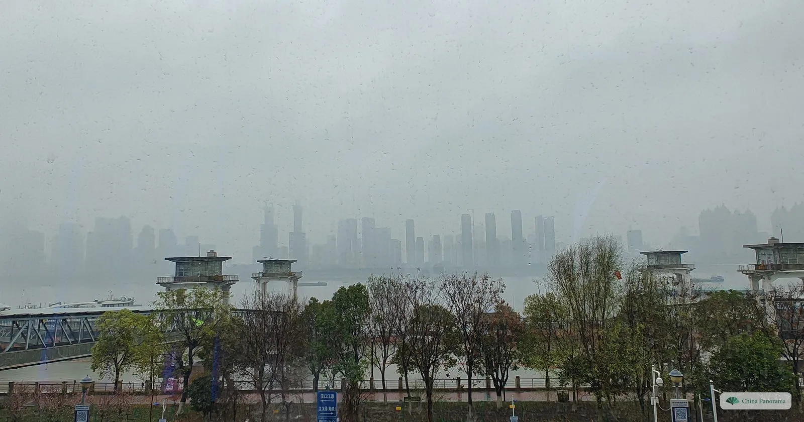 View from a terrace over trees and old pier buildings to a wide river and a skyline of towers dissolving in rain and fog