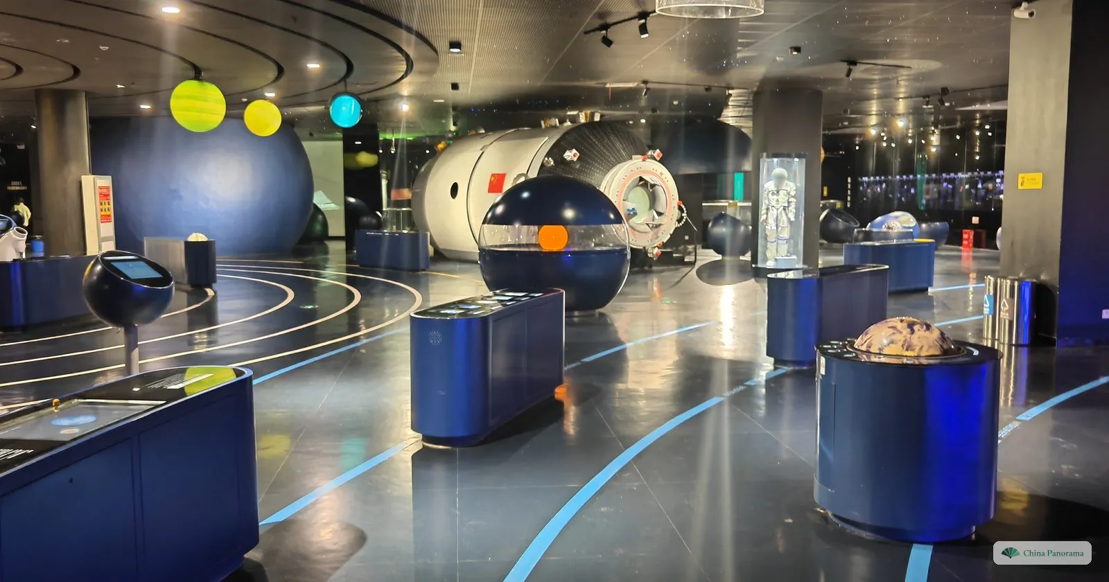 A dark exhibition hall with a model space station, coloured planets, an astronaut suit in a case and display pedestals
