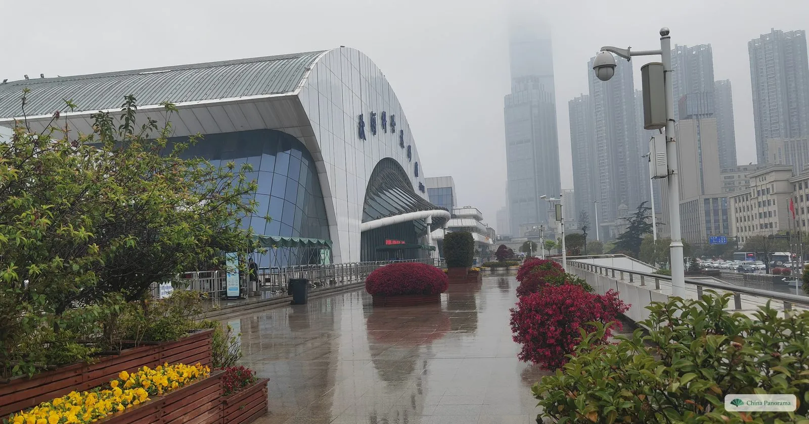 A large building with a curved silver roof beside a rainy riverside plaza with flower beds, skyscrapers in mist behind