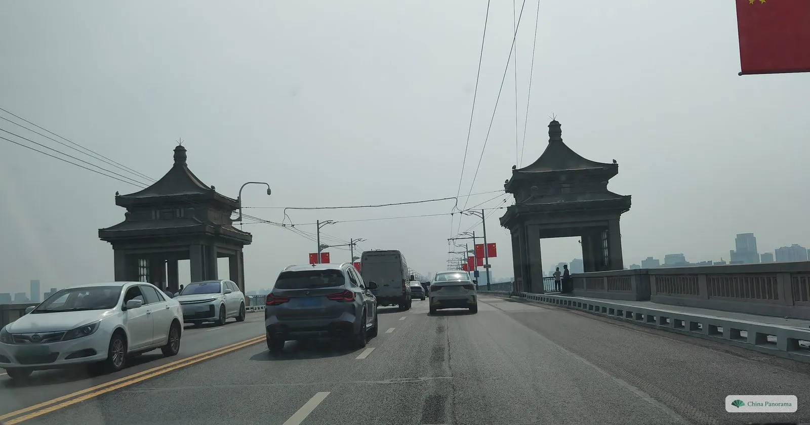 Seen through a car windscreen: a bridge road with cars, and two dark stone pavilions with upturned roofs standing either side of it, trolleybus wires overhead