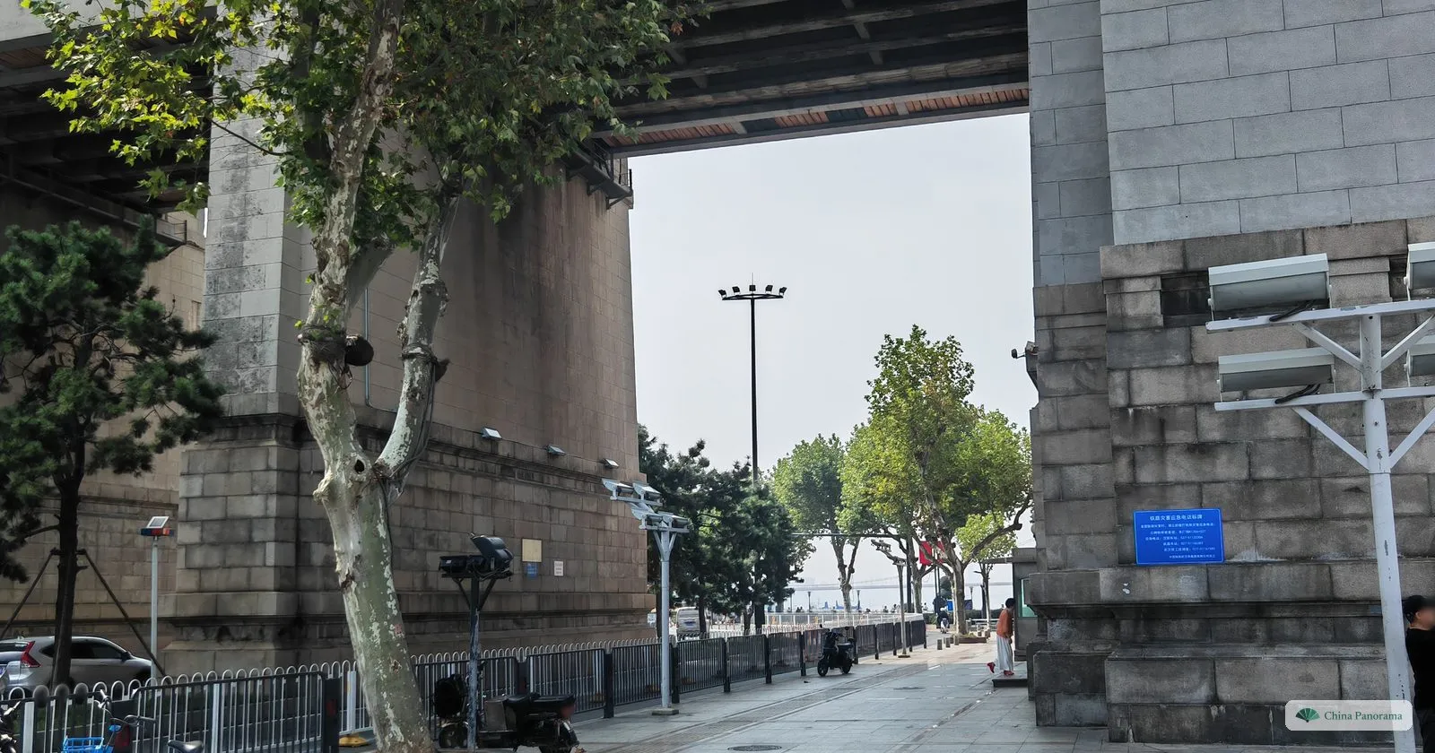 A riverside lane passing under the deck of a bridge between huge granite pillars, plane trees and street lamps