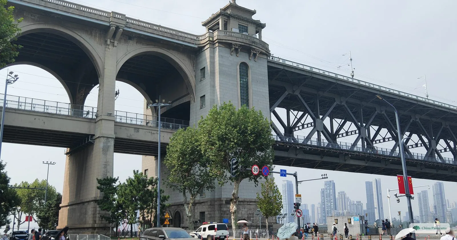 A tall stone tower with a small pavilion on top at the end of a steel truss bridge, a road with traffic lights passing under great stone arches, skyscrapers beyond