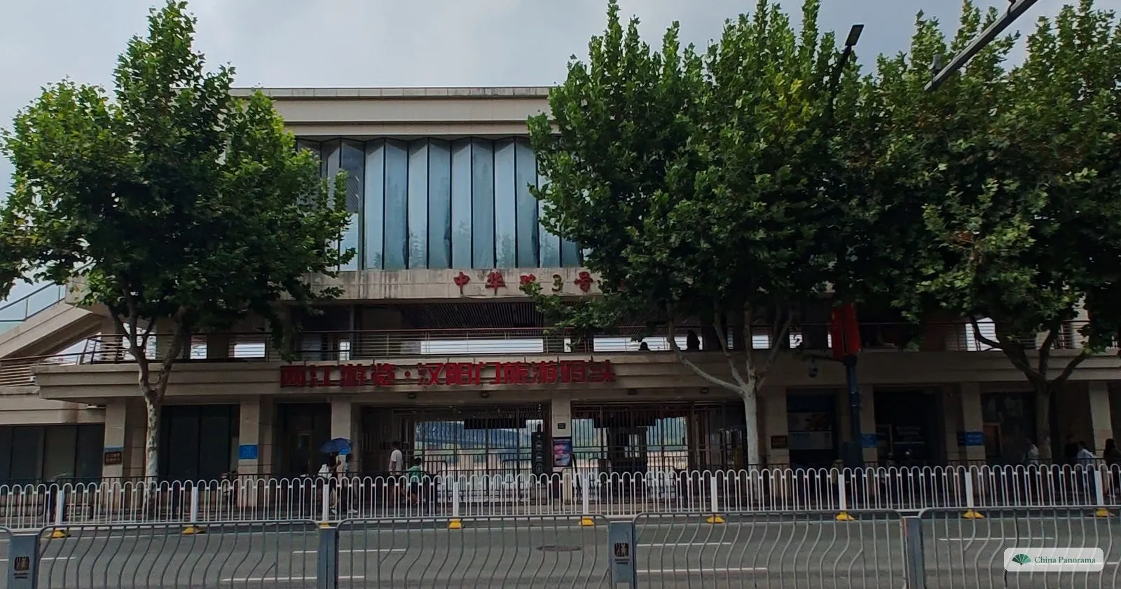 A two-storey ferry terminal with a red sign behind plane trees and a long metal railing along the road