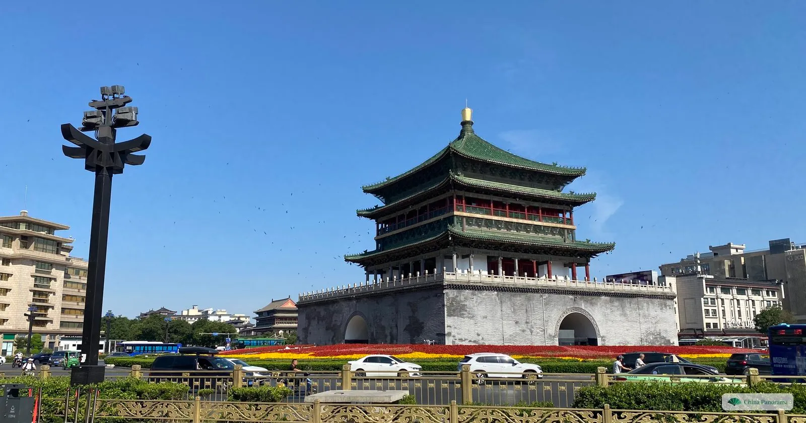 The Bell Tower of Xi’an, a three-eaved green-roofed wooden tower on a square brick base with an archway, in the middle of a roundabout planted with flowers, under a clear blue sky