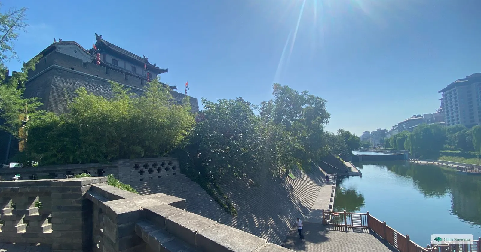 The moat of Xi’an beside the city wall, with a brick parapet in the foreground, a tower on the wall and a riverside path in the low sun