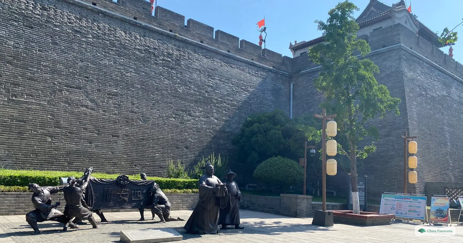The grey-brick city wall of Xi’an at full height, with battlements and a tower on top and a group of bronze figures on the pavement at its foot