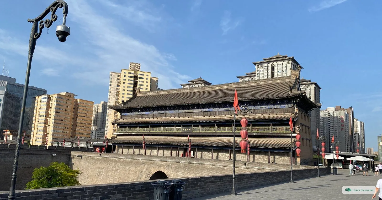 A three-tiered gate tower hung with red lanterns beside a wide brick-paved walkway with a parapet, and apartment blocks behind