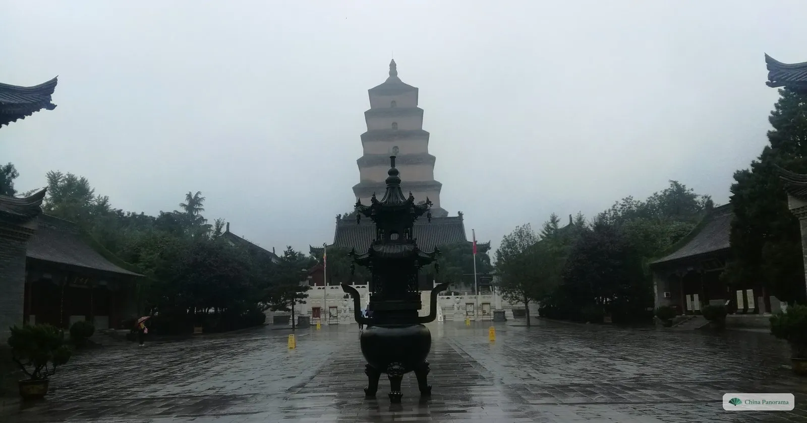 A bronze incense burner in a rain-wet courtyard of Daci’en Temple, with the Giant Wild Goose Pagoda rising behind it into low cloud