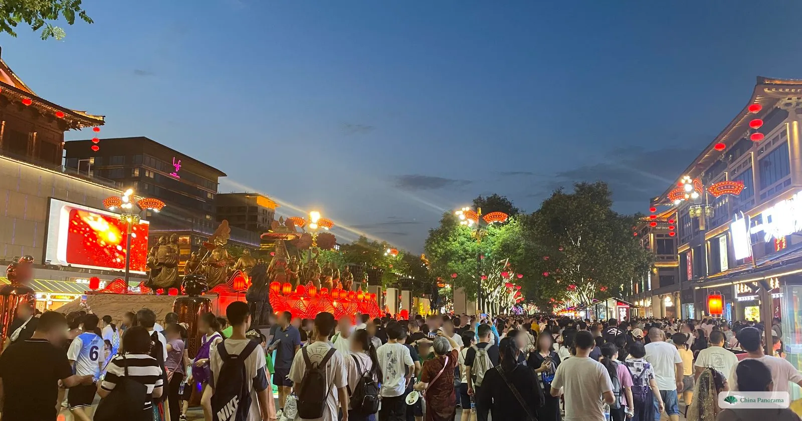 A dense crowd walking down Grand Tang Mall at dusk between lit Tang-style buildings, with a row of sculptures and red lanterns on the left