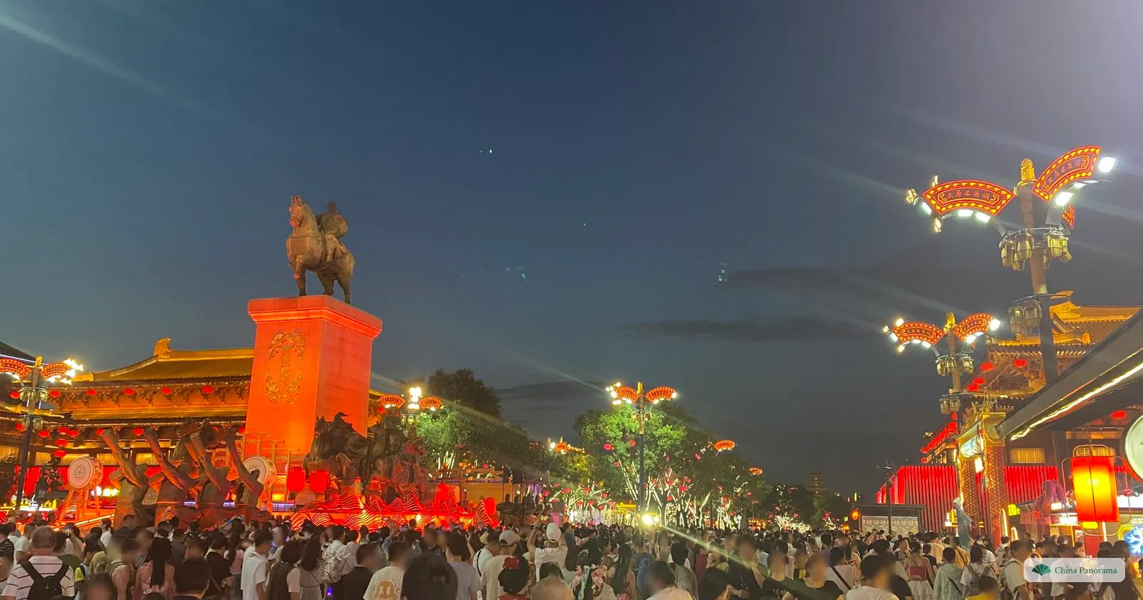 An equestrian statue on a tall column lit red above a packed crowd in Grand Tang Mall after dark, with lit Tang-style halls and lamp standards on either side